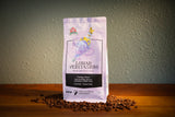 A bag of Treasured Grounds 'Lunar Veritasium' coffee stands upright on a wooden surface with scattered coffee beans around its base. The packaging is light lavender with black and white text, and the background is a textured gray wall.