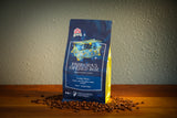 A bag of coffee stands upright on a wooden surface with scattered coffee beans around its base. The package is dark blue with gold and white graphics and text reading 'Pandora's Opened Box' and 'Medium Roast Coffee.